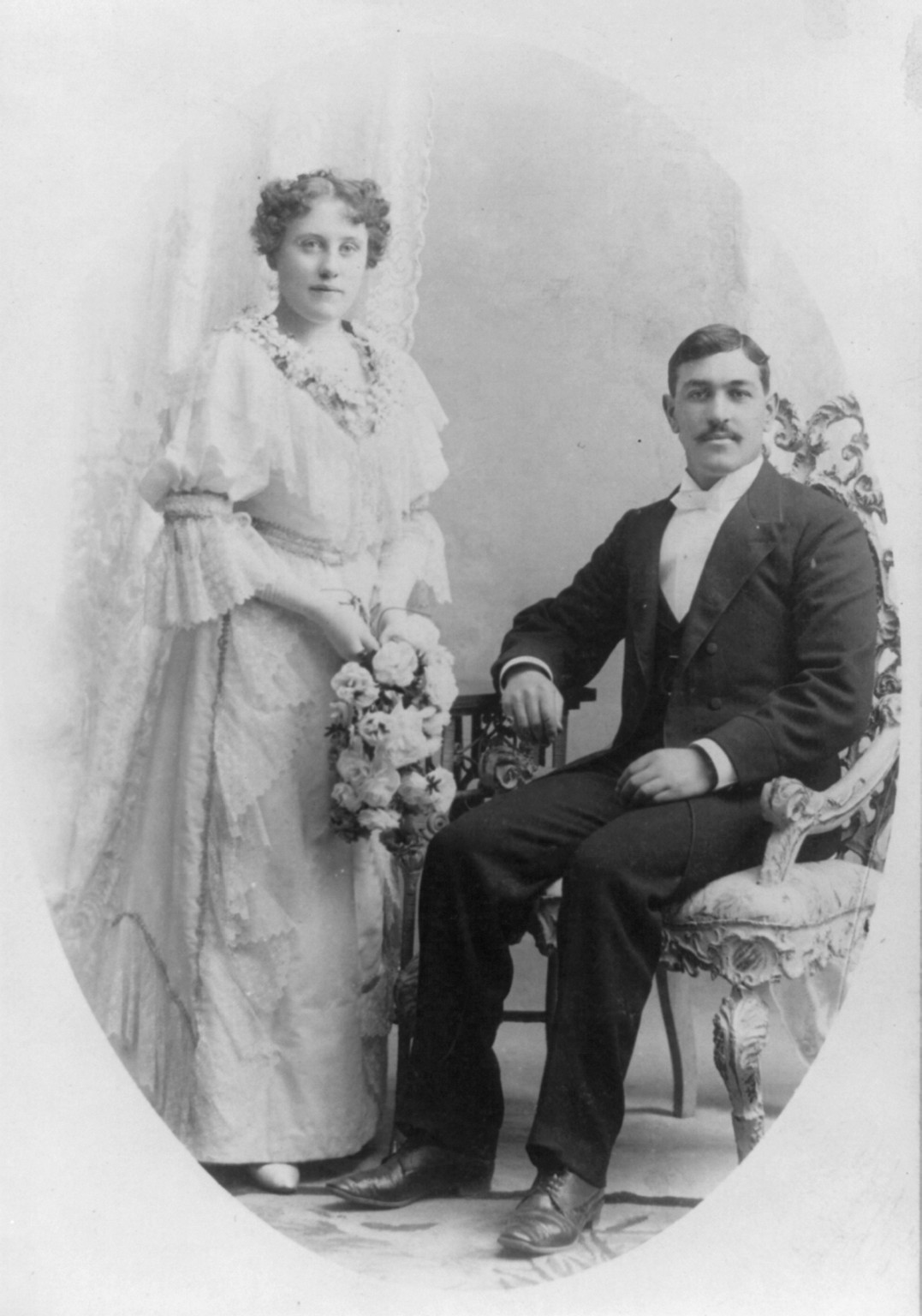 A woman holding flowers stands beside a seated man in an unidentified wedding portrait